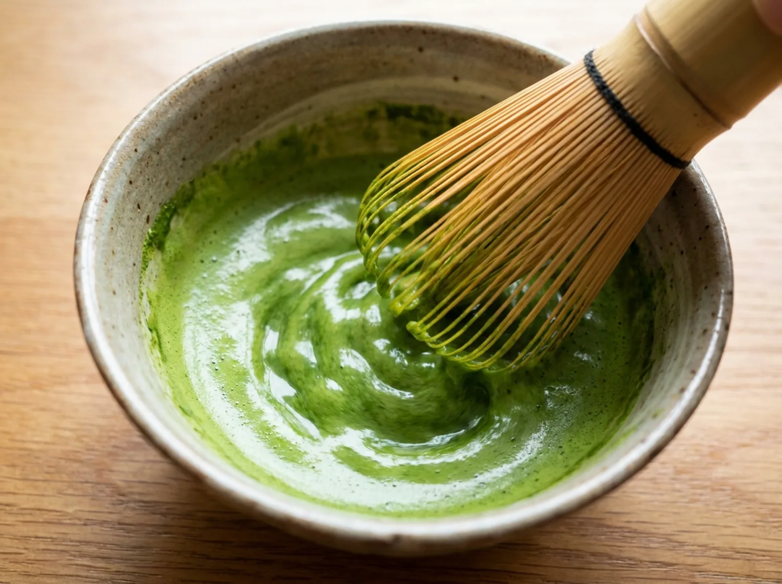 A bamboo chasen whisking bright green matcha and hot water into a smooth paste in a bowl, before any milk is added