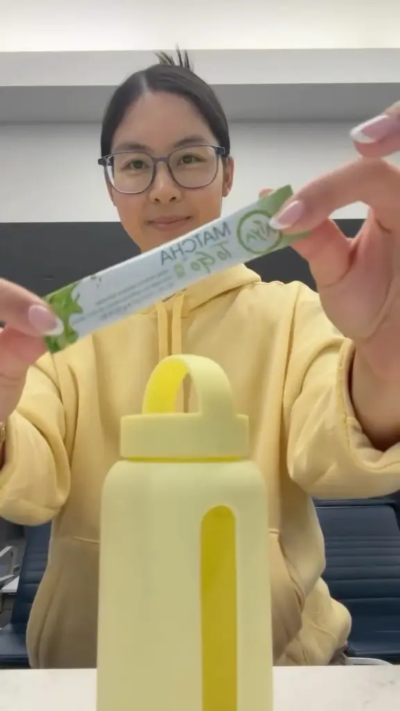 Person in a yellow hoodie holding a Matcha To Go packet above a yellow bottle