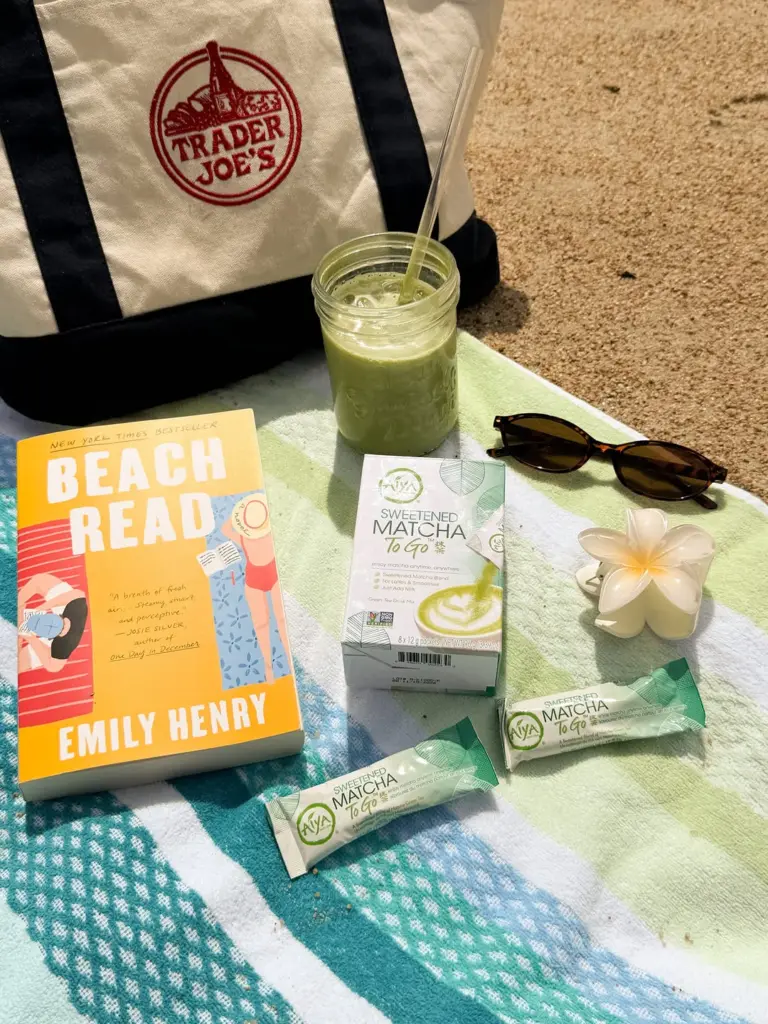 Iced green matcha drink, Aiya packets, a book, and sunglasses arranged on a beach towel
