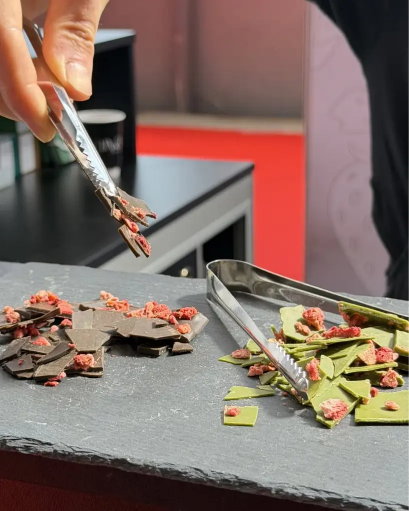 Tongs lifting dark chocolate pieces beside green and dark chocolate topped with red crumbs