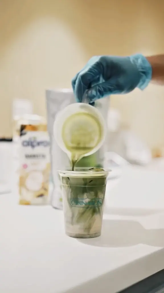 Gloved hand pouring a green drink into a clear cup filled with ice and milk