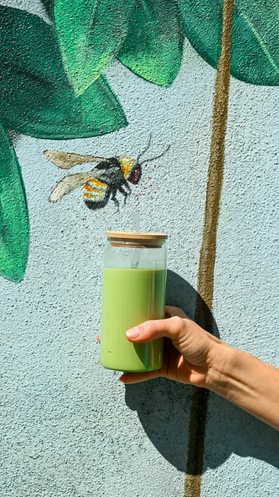 Hand holding a glass of pale green iced drink against a painted bee mural