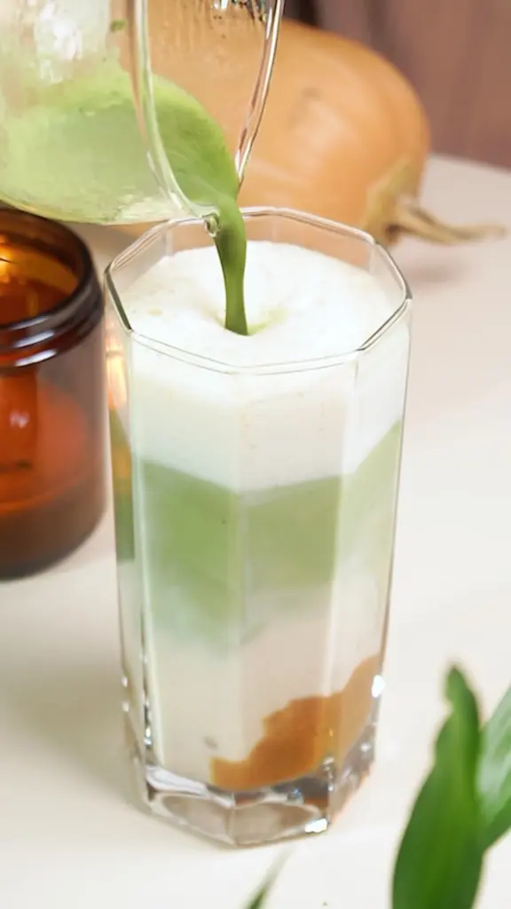 Green liquid being poured into a tall glass filled with a white foamy drink