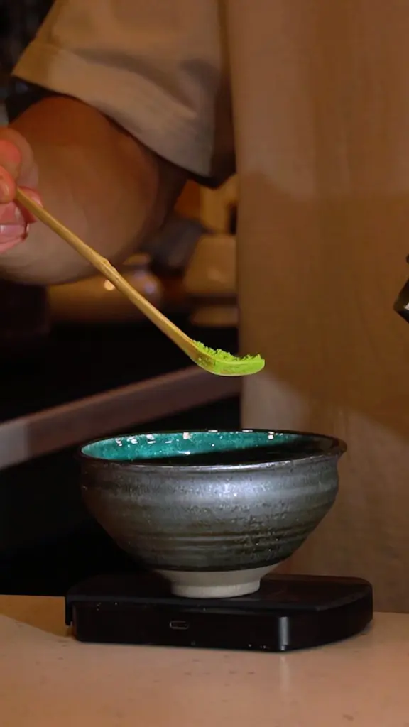 Bamboo scoop holding bright green powder above a ceramic bowl on a scale