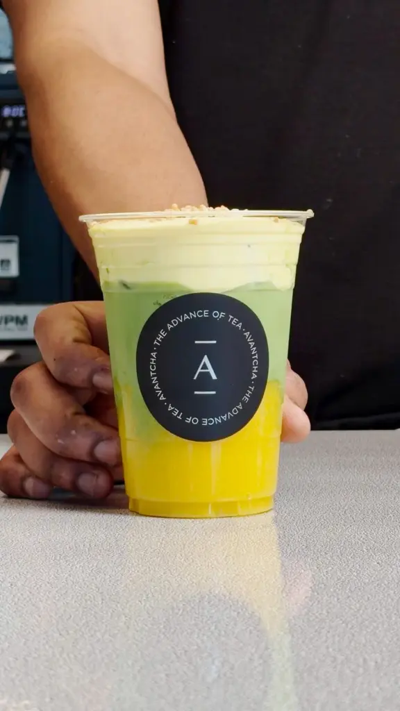Hand holding a clear cup with yellow and green layers beneath a thick pale topping