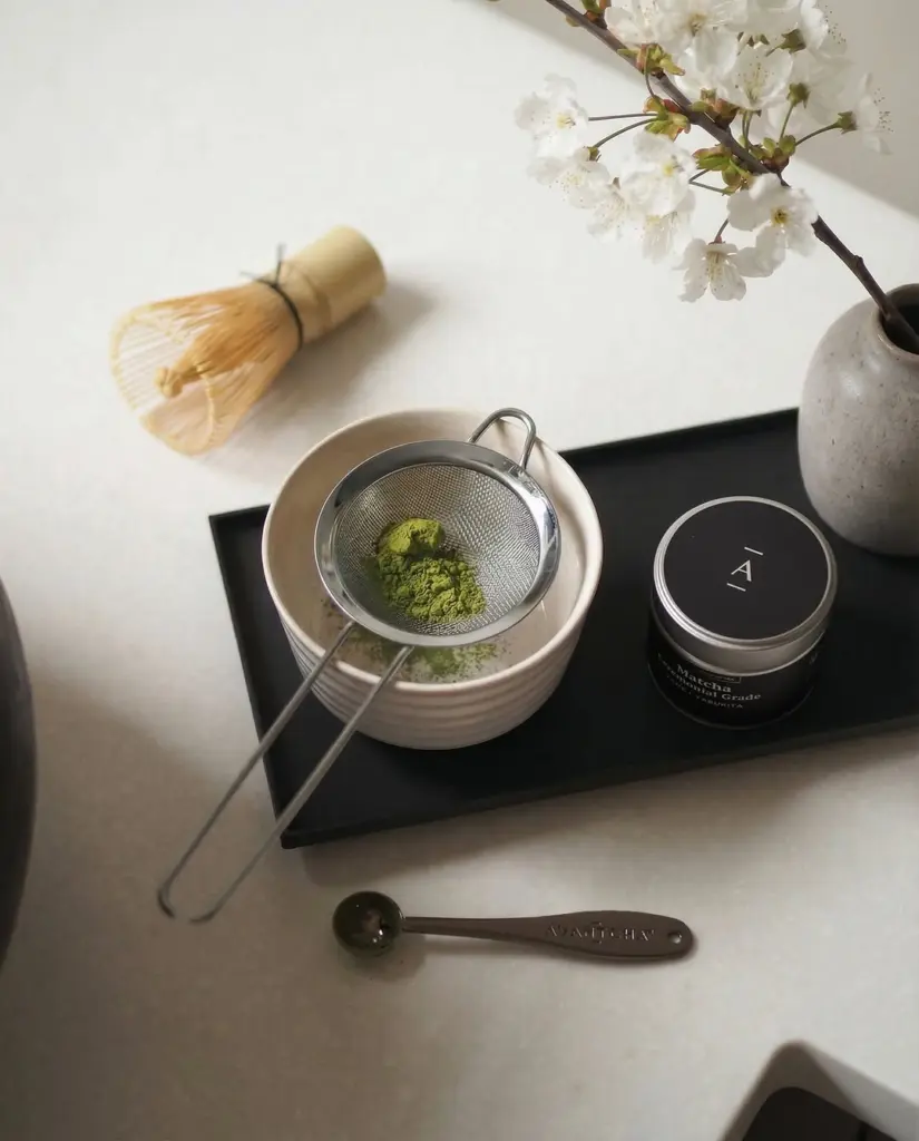 Green matcha powder in a small sieve over a bowl beside a tin, bamboo whisk, spoon, and white blossoms