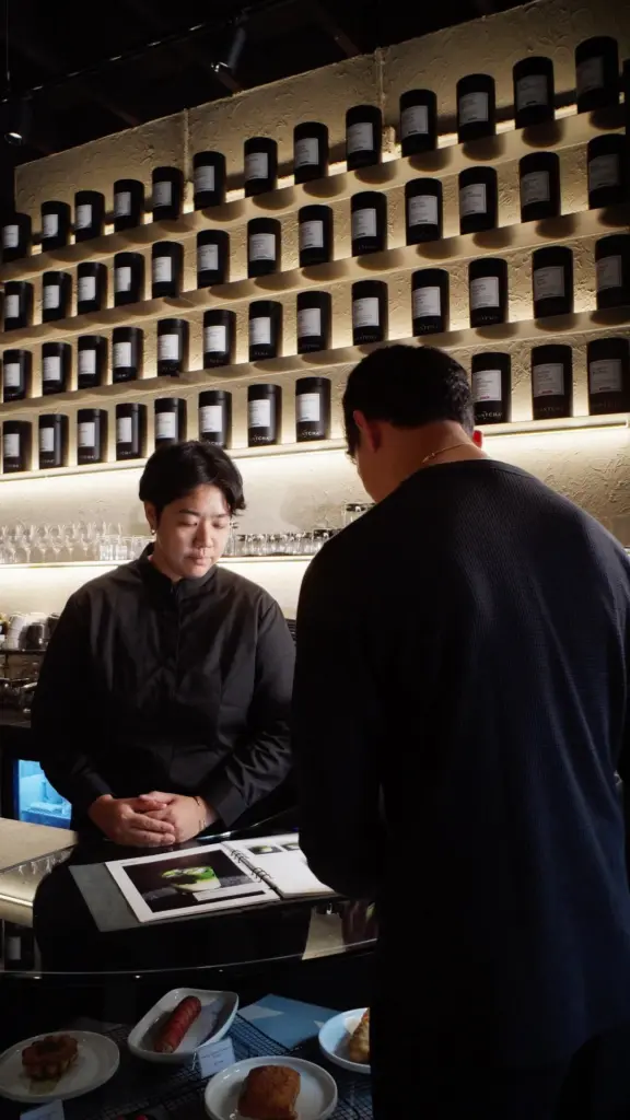Two people at a counter beneath illuminated shelves of black tea canisters