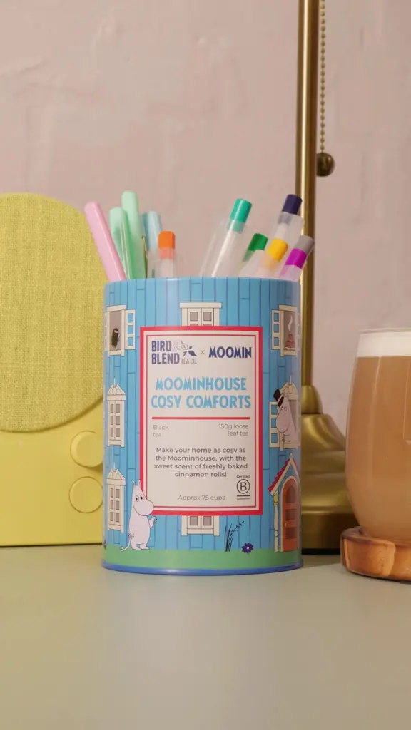 Blue Moominhouse Cosy Comforts tea tin holding colorful pens beside a glass of milky tea