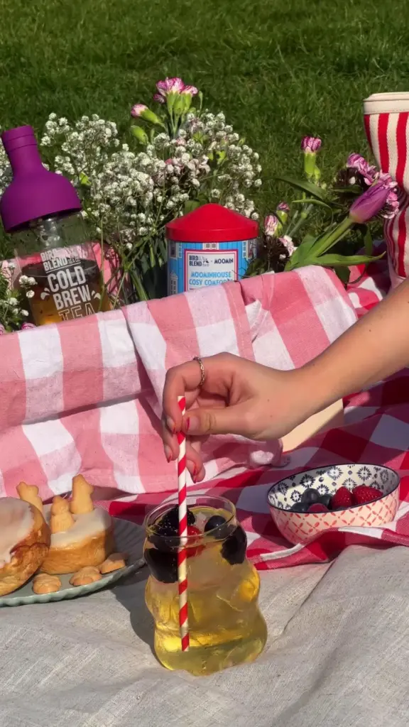 Hand stirring a fruit-filled iced tea beside pastries, berries, flowers, and tea containers on a picnic blanket
