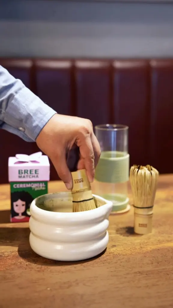 Hand whisking green matcha in a white bowl with a bamboo whisk
