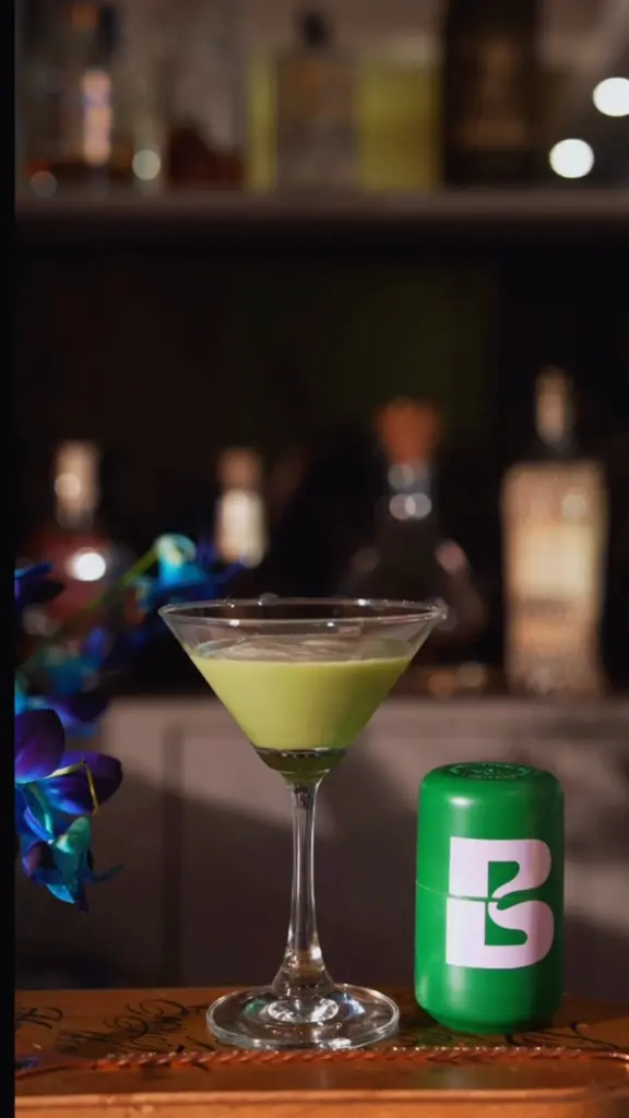 Pale green drink in a martini glass beside a green branded container