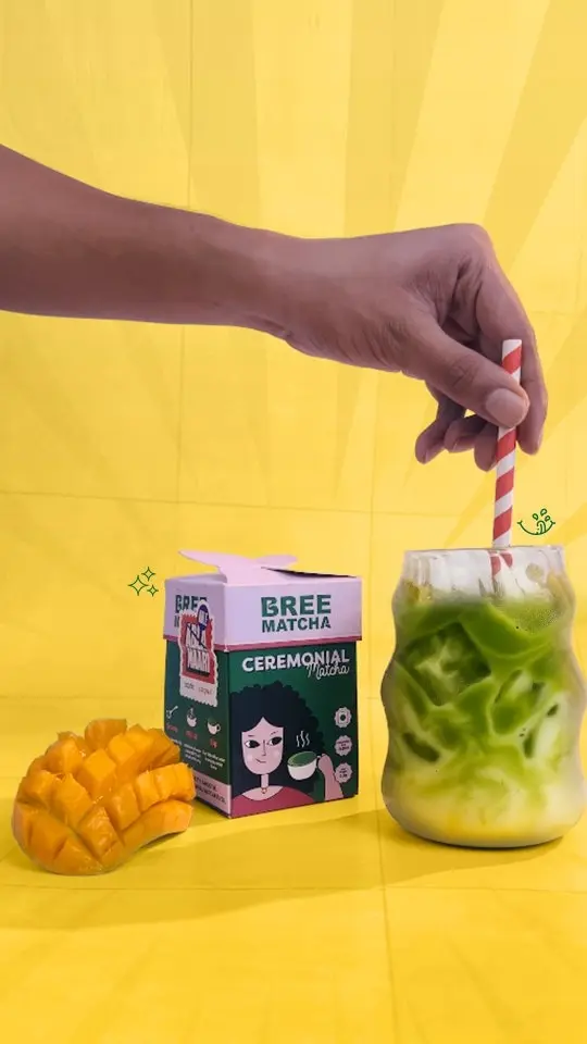 Hand stirring an iced green drink beside Bree Matcha packaging and sliced mango