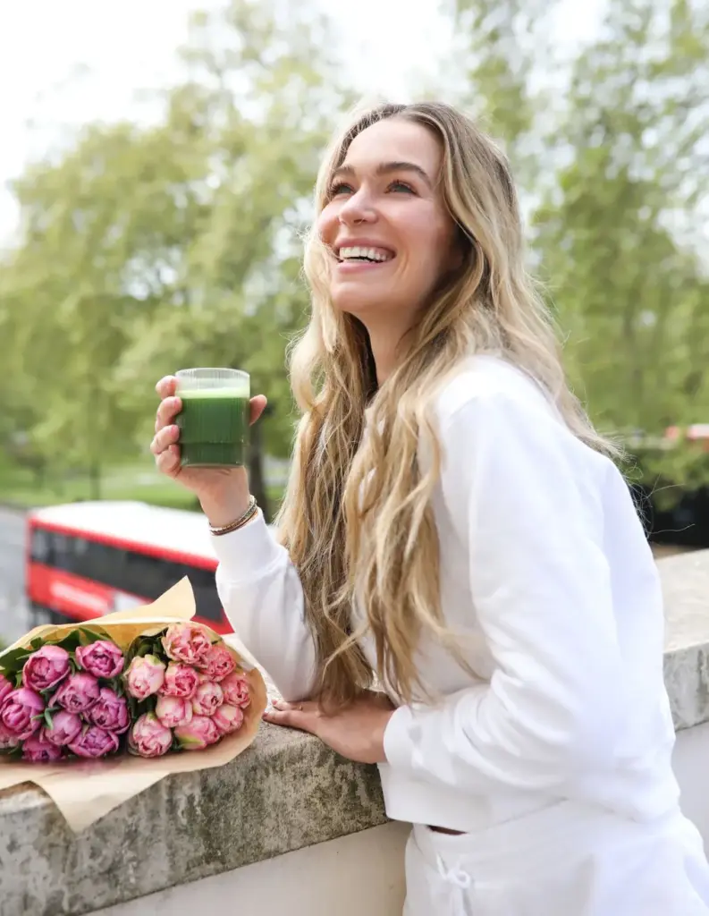 Smiling woman in white holding a glass of green drink beside a bouquet of pink flowers outdoors