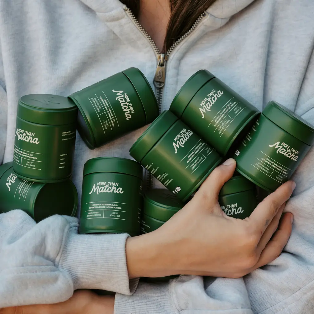 Person in a gray hoodie holding several dark green More Than Matcha ceremonial matcha powder tins