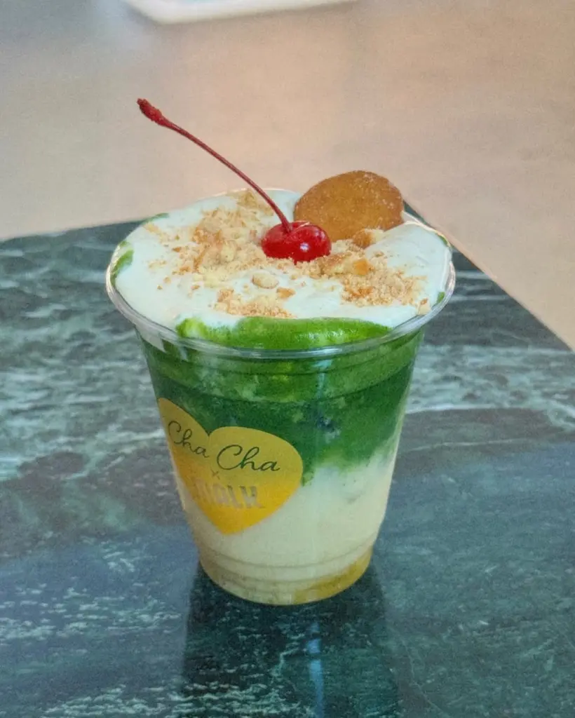 Layered green drink topped with foam, a cherry, cookie, and crumbs