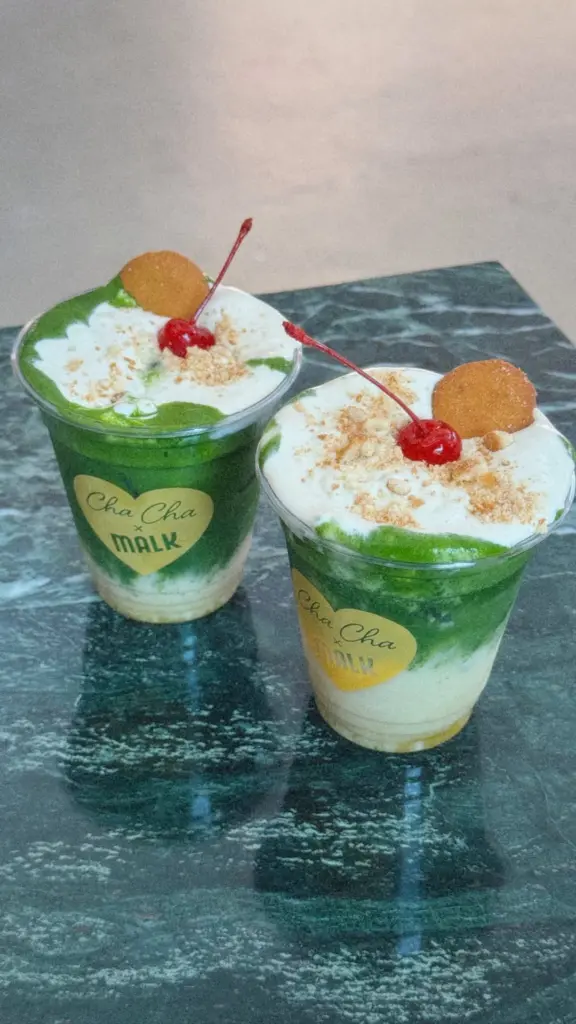 Two green layered drinks topped with foam, cherries, cookies, and crumbs