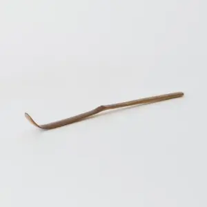 Slender brown bamboo matcha scoop with a curved tip on a white surface