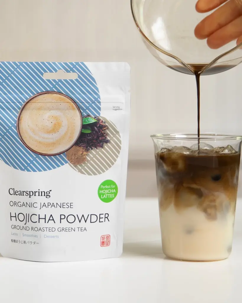 Hojicha powder pouch beside an iced latte as dark liquid is poured into the glass