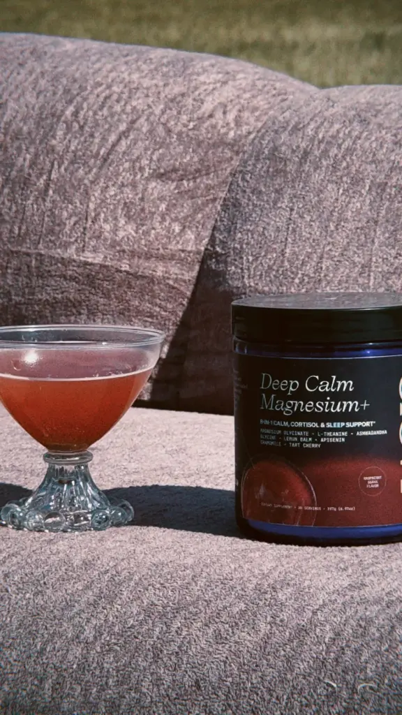 Deep Calm Magnesium+ container beside a glass of reddish-brown liquid on a textured chair