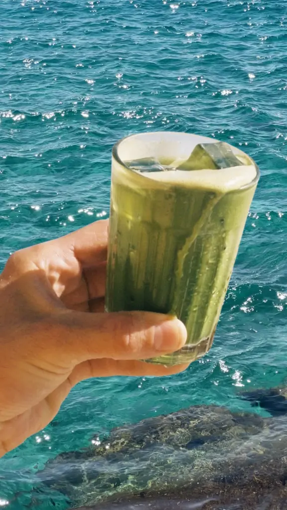 Hand holding a foamy iced green drink above sunlit turquoise water