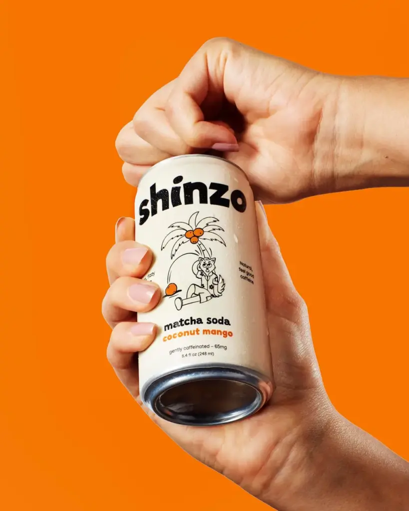 Hands opening a can of Shinzo coconut mango matcha soda against an orange background