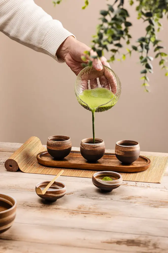 Bamboo chashaku resting on a small bowl as green matcha is poured into one of three cups