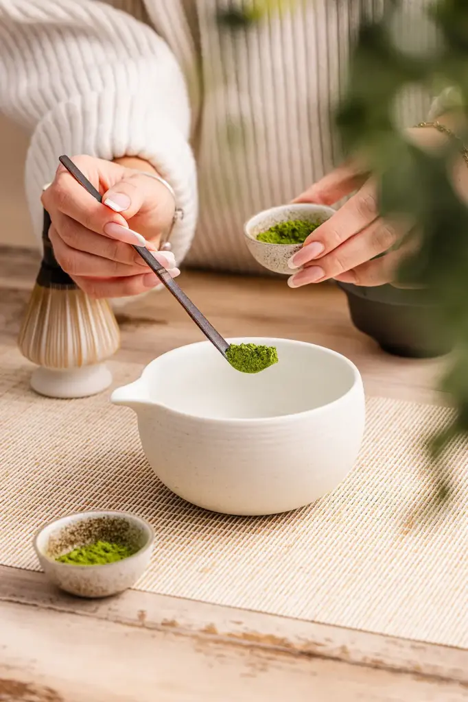 Dark bamboo chashaku holding green matcha powder above a white preparation bowl