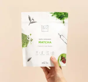 Hand holding a white Matcha & CO culinary matcha powder pouch against a pale peach background