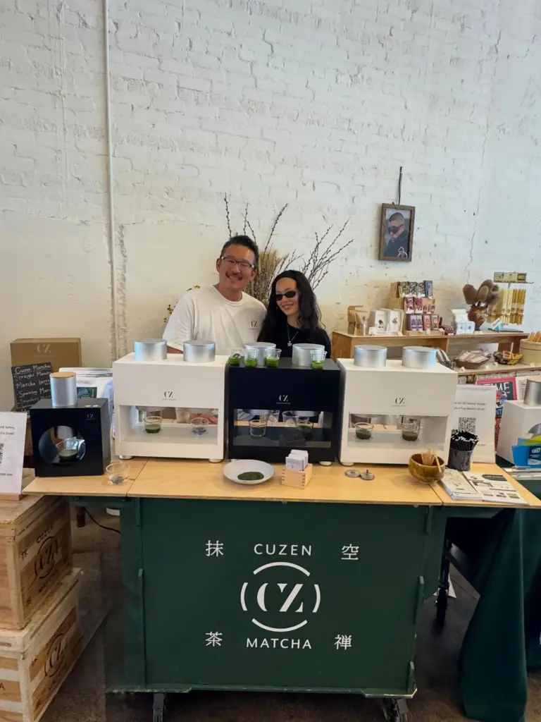 Two people standing behind a Cuzen Matcha counter with several machines