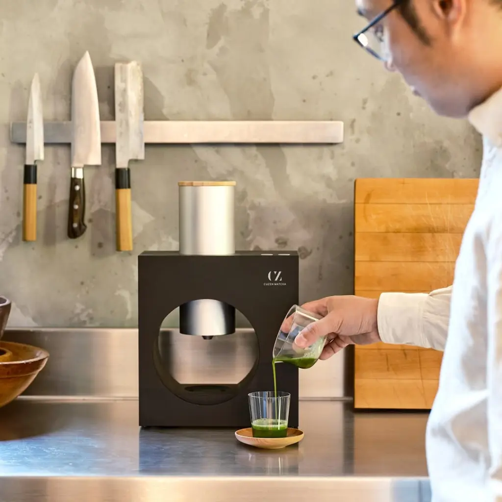 Person pouring bright green tea into a small glass beside a black Cuzen Matcha machine