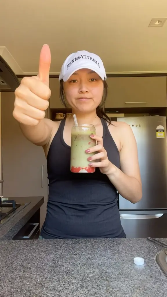 Person in a kitchen holding a layered green iced drink and giving a thumbs-up
