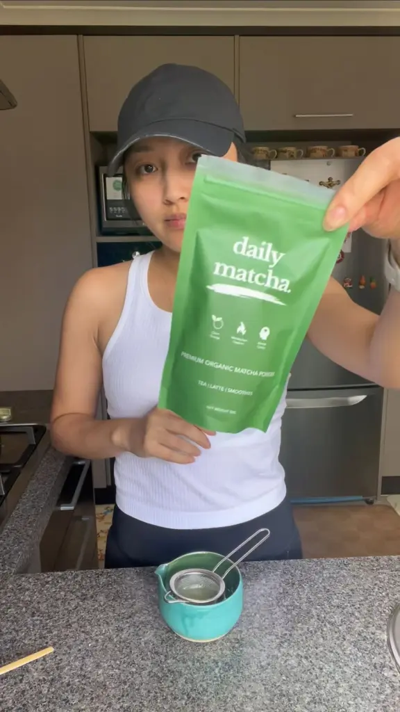 Person holding a green Daily Matcha pouch above a bowl and fine-mesh strainer