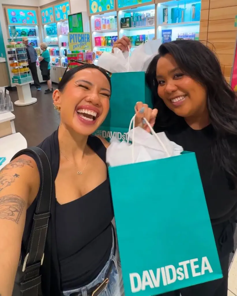 Two smiling shoppers holding turquoise David's Tea bags inside a colorful store