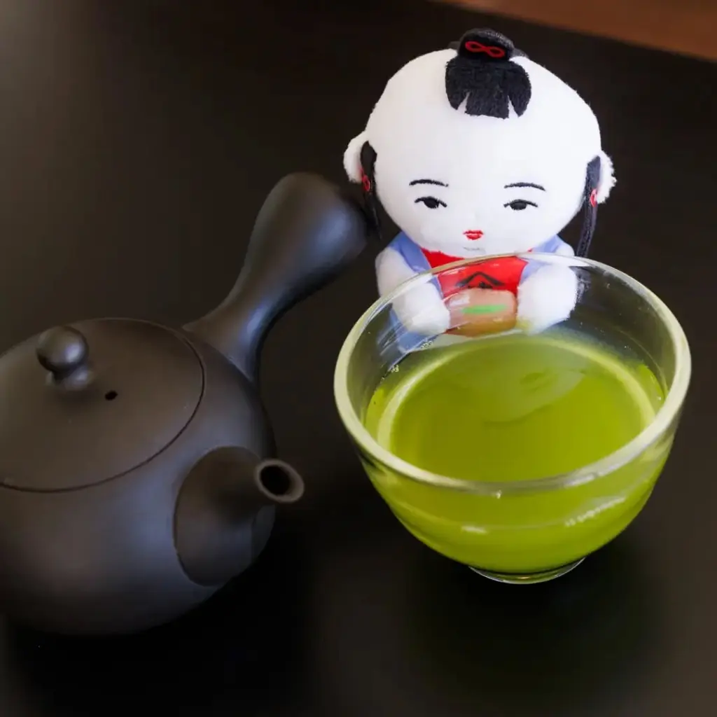 Glass cup of bright green tea beside a black teapot and small decorative doll