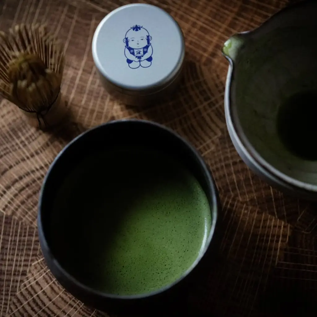 Bowl of frothy green matcha beside a bamboo whisk, lidded container, and pitcher