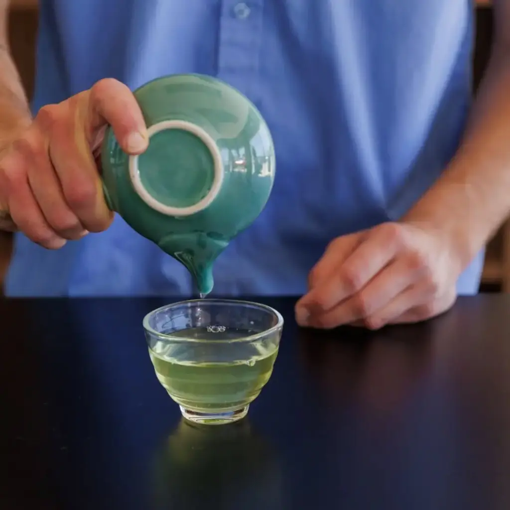 Hands pouring pale green tea from a turquoise cup into a small glass cup