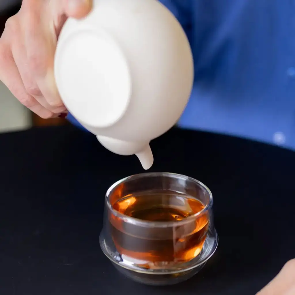 Hand holding a white teapot above a clear glass cup of amber tea