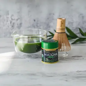 Aprika Life matcha tin beside a glass bowl of green tea and bamboo whisk