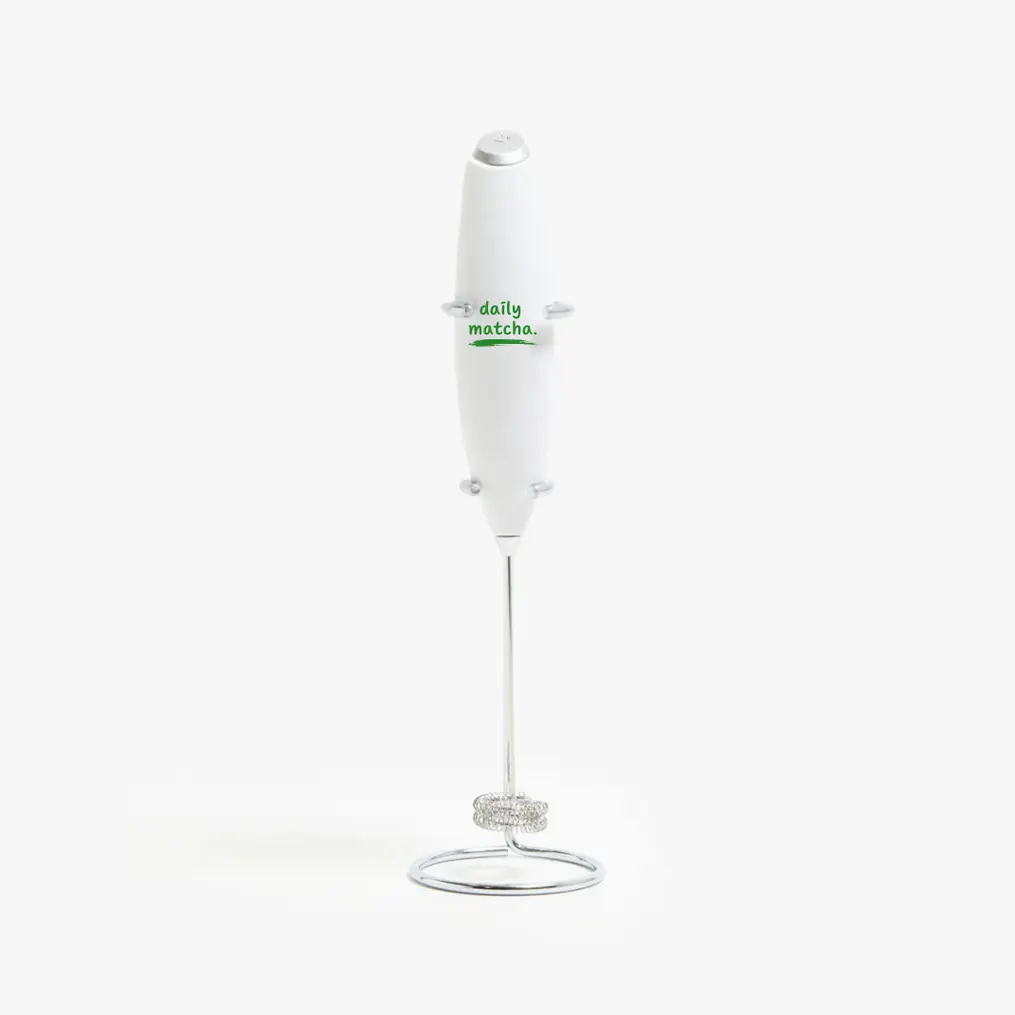 White handheld electric matcha whisk with green Daily Matcha logo, resting upright in a chrome wire stand