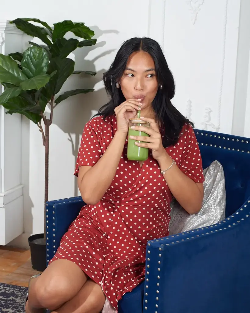 Woman in a red polka-dot dress sipping an iced green matcha drink in a blue armchair