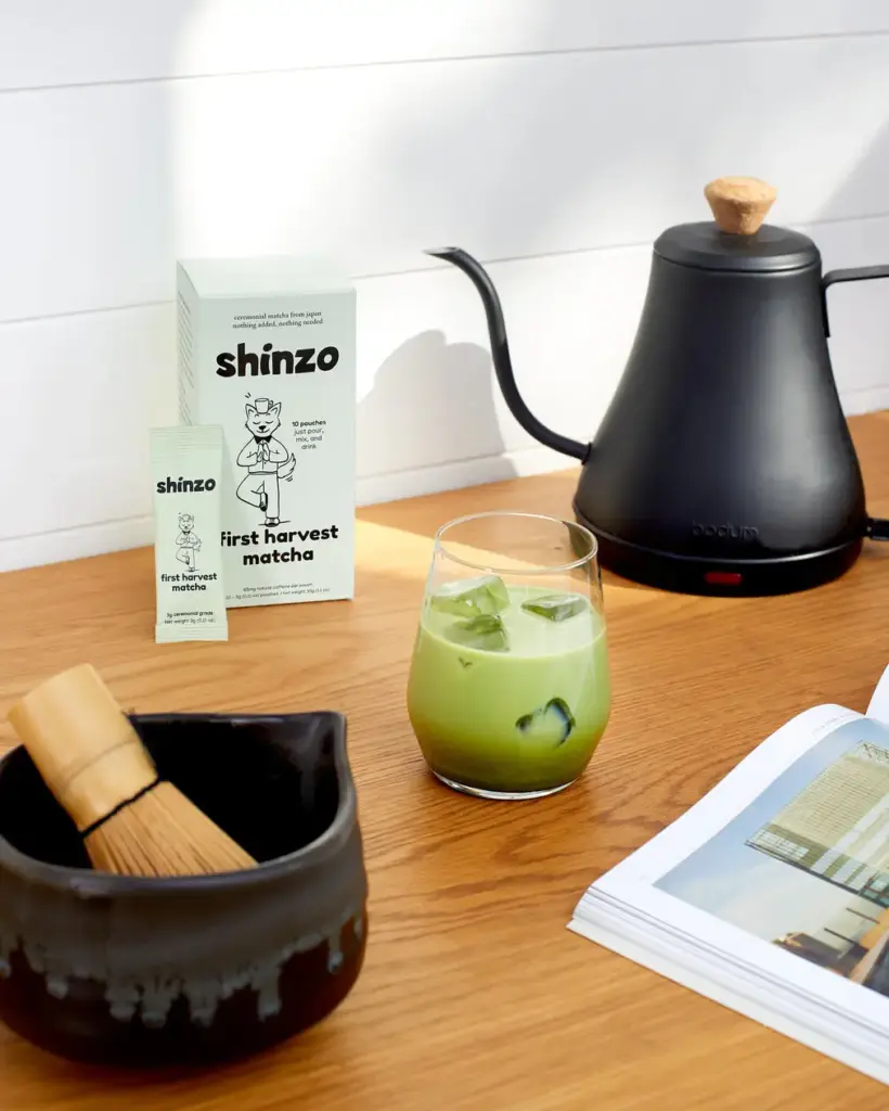 Shinzo First Harvest Matcha box and pouch beside an iced green drink, kettle, bowl, and bamboo whisk