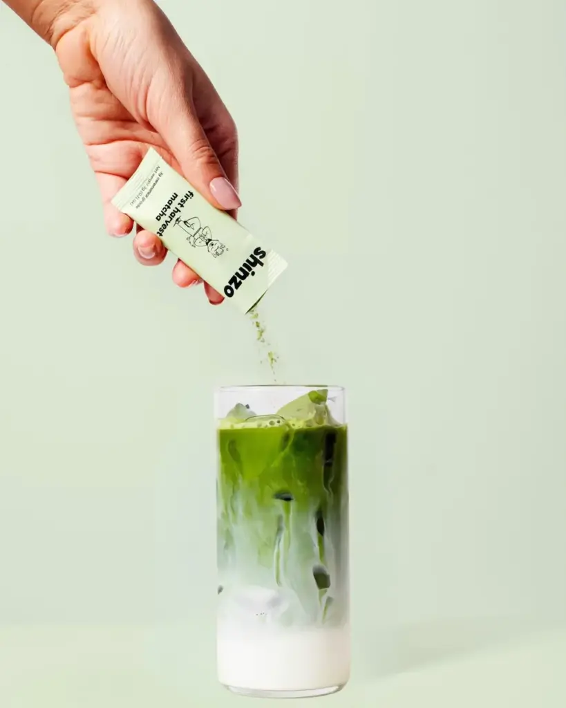 Hand pouring Shinzo First Harvest Matcha powder from a sachet into an iced green and white drink