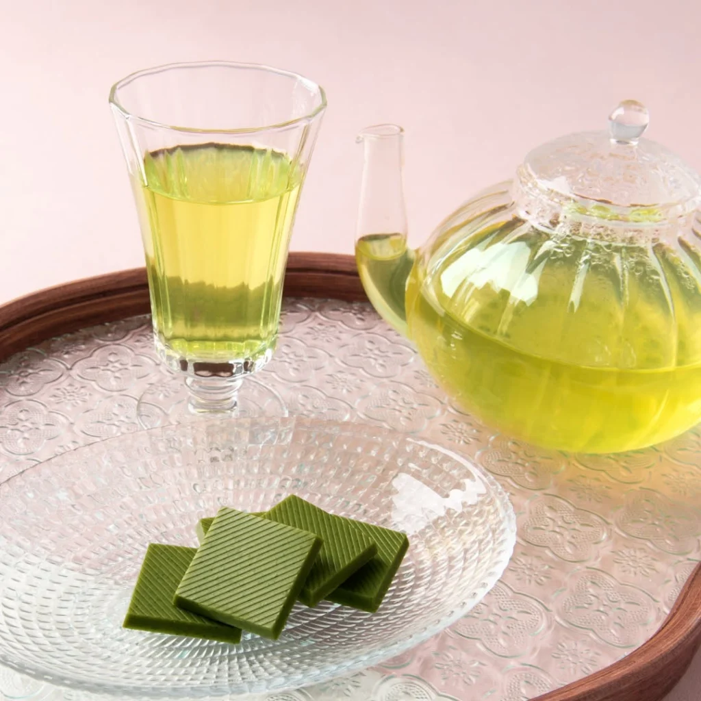 Glass teapot and cup of pale green tea beside square green confections