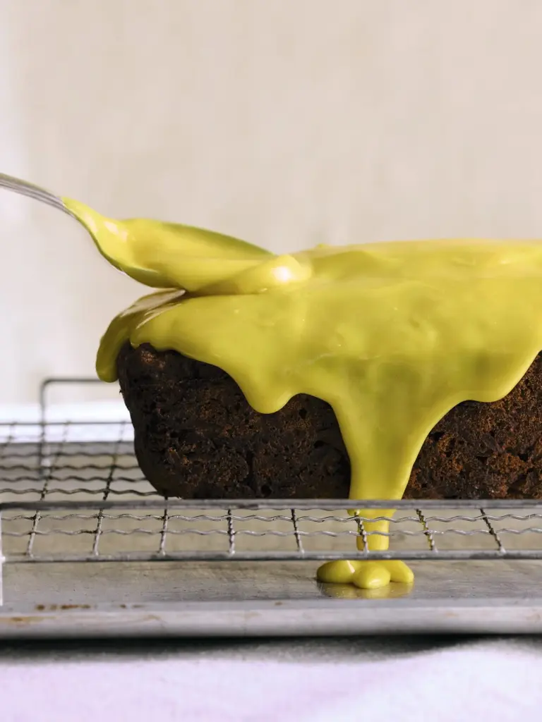 Green glaze being spooned over a dark loaf cake on a cooling rack