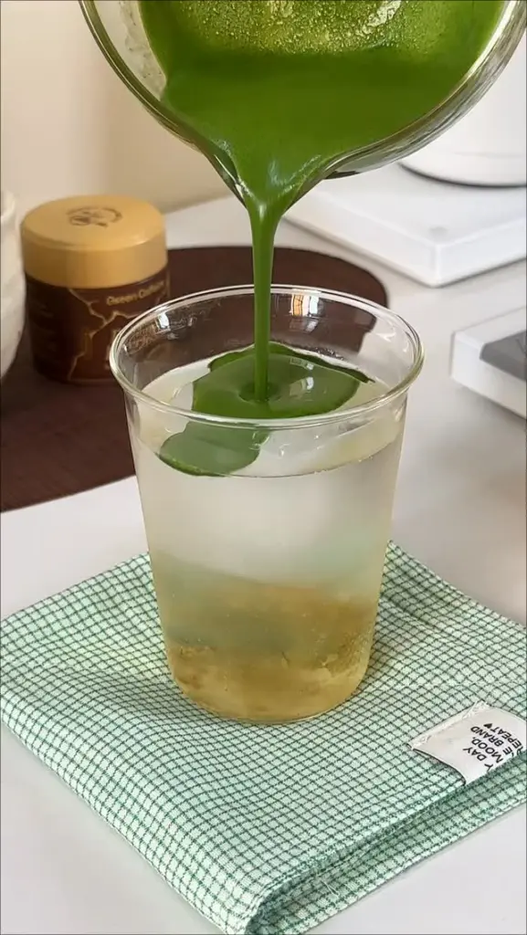 Thick green matcha pouring into a clear cup filled with ice and a pale layered drink