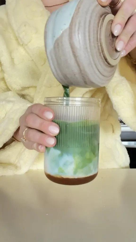 Hands pouring a green drink from a ceramic bowl into a ribbed glass with ice and milk