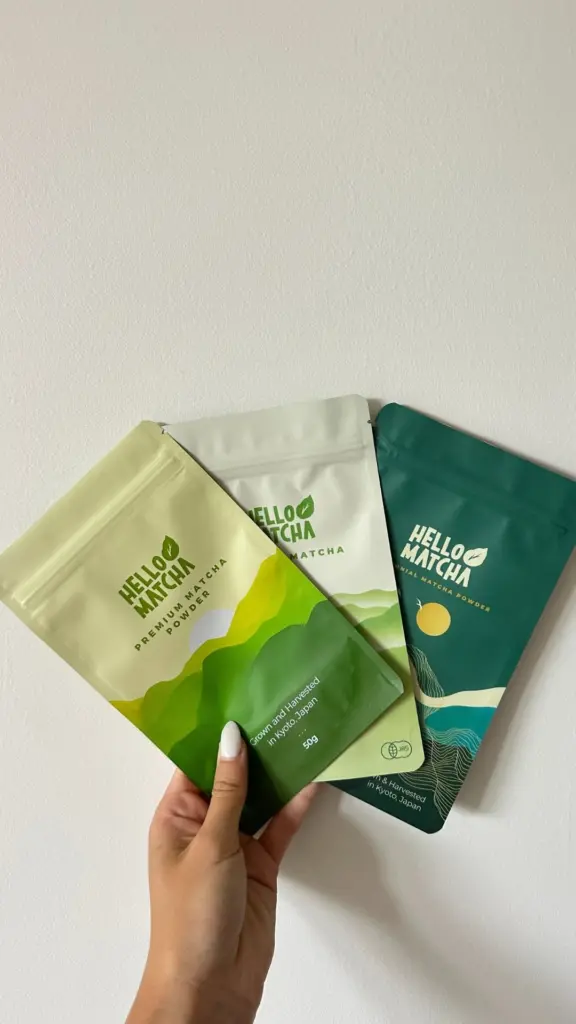 Hand holding three colorful Hello Matcha powder pouches against a white wall