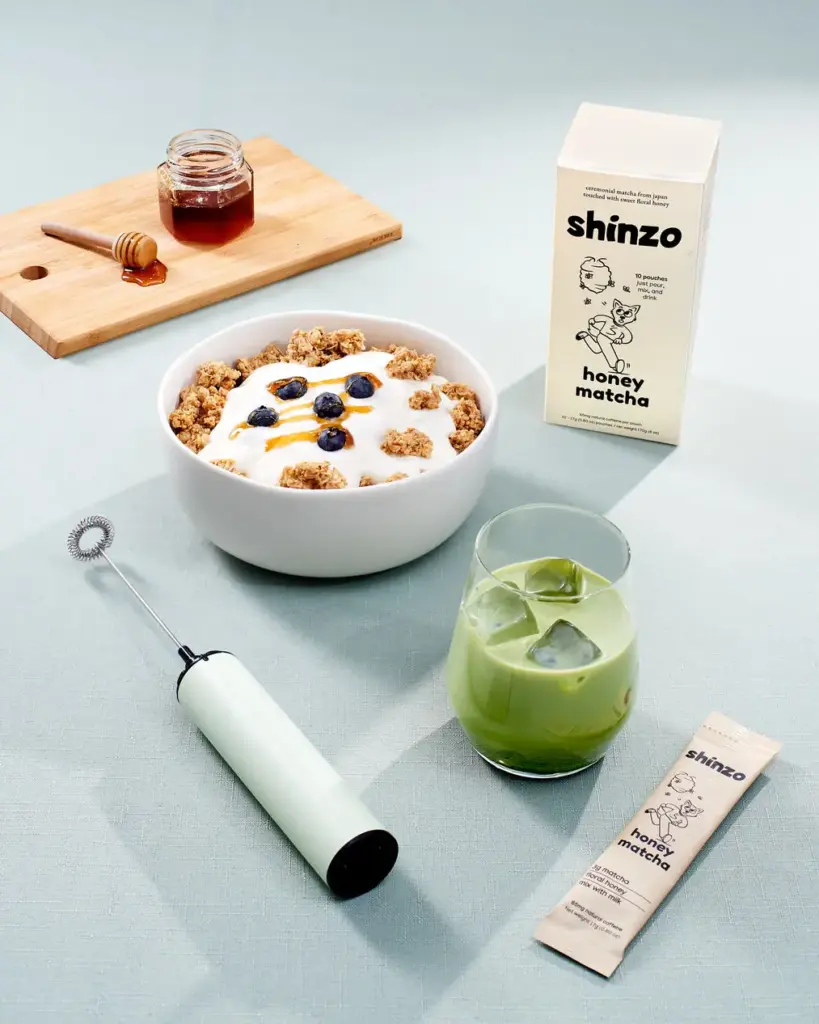 Shinzo Honey Matcha box and pouch beside an iced green drink, frother, yogurt bowl, and jar of honey