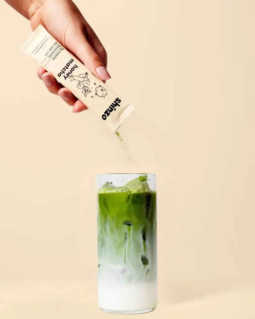 Hand pouring Shinzo Honey Matcha powder from a single-serve pouch into a layered iced green drink