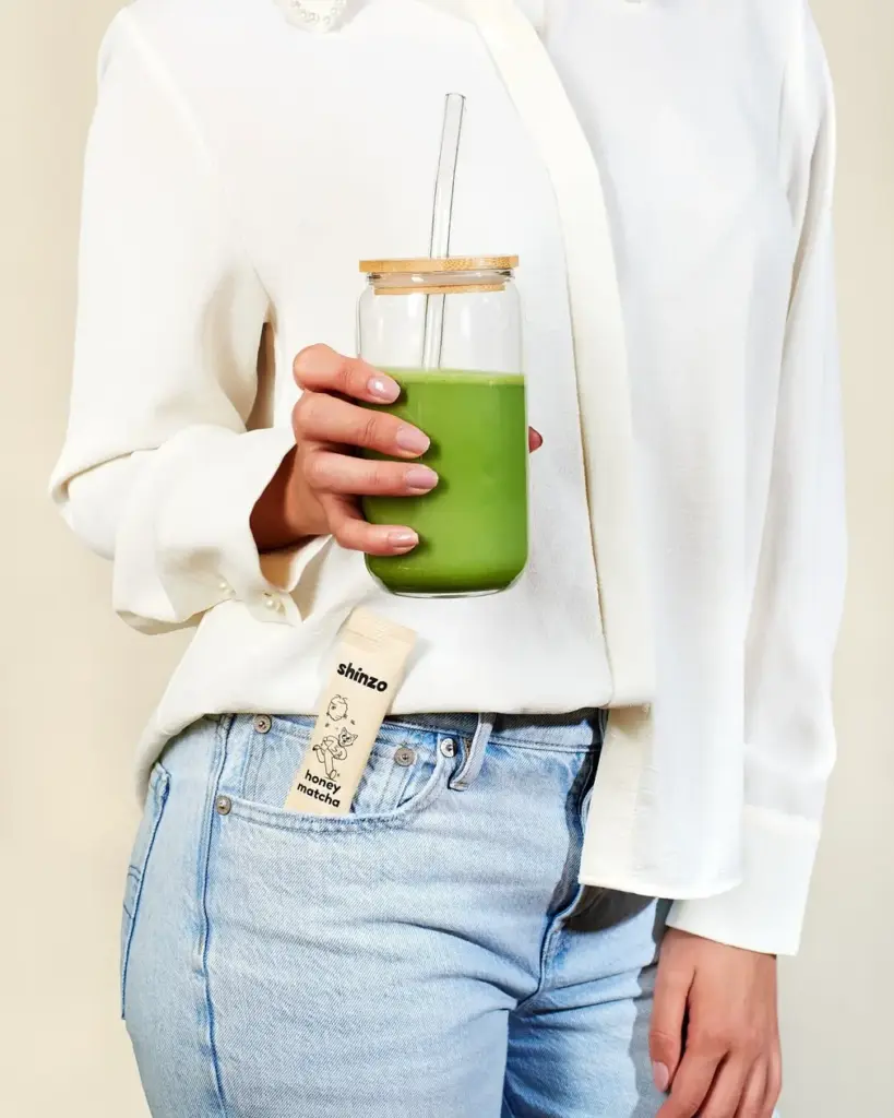 Person holding a glass of green matcha with a Shinzo Honey Matcha pouch in a jeans pocket